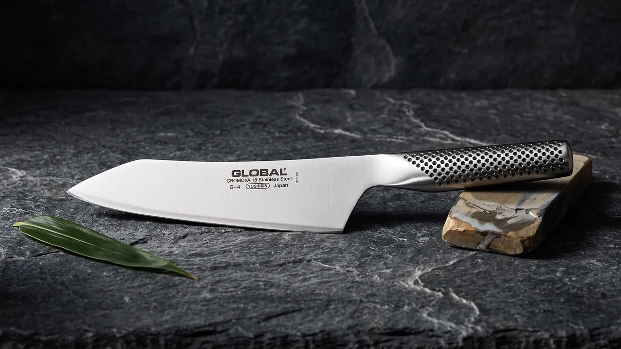 Global G-4 oriental de 18 cm sobre pizarra oscura rugosa, junto a un fragmento de piedra de afilar y una hoja de bambú