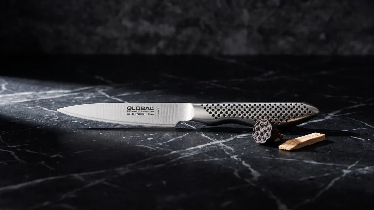 Global GS-38 pelador de 9 cm sobre mármol negro con vetas grises, junto a una semilla de loto y una esquirla de madera de hinoki