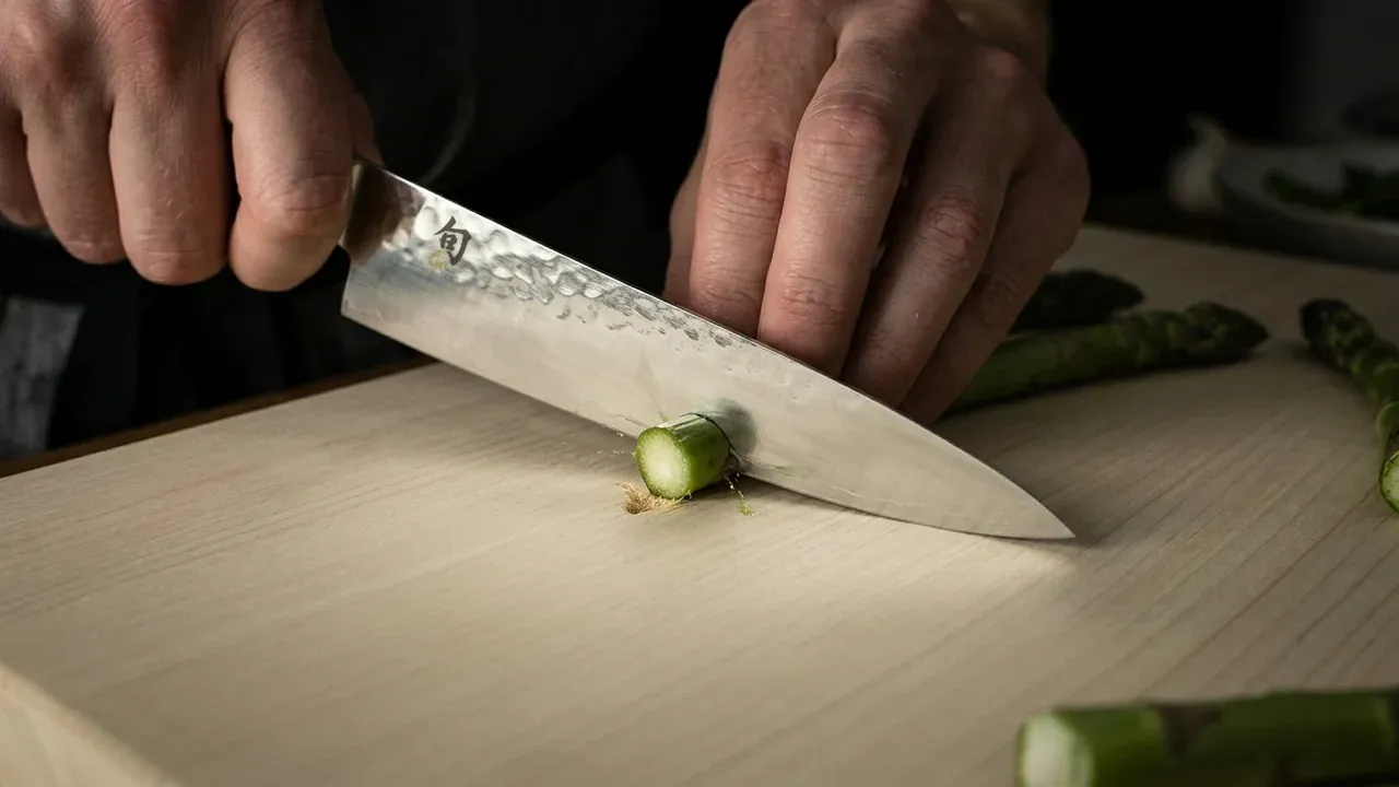 Cuchillo japonés cortando un espárrago sobre la tabla de hinoki KAI Shun, con el filo hundiéndose en la madera blanda de ciprés