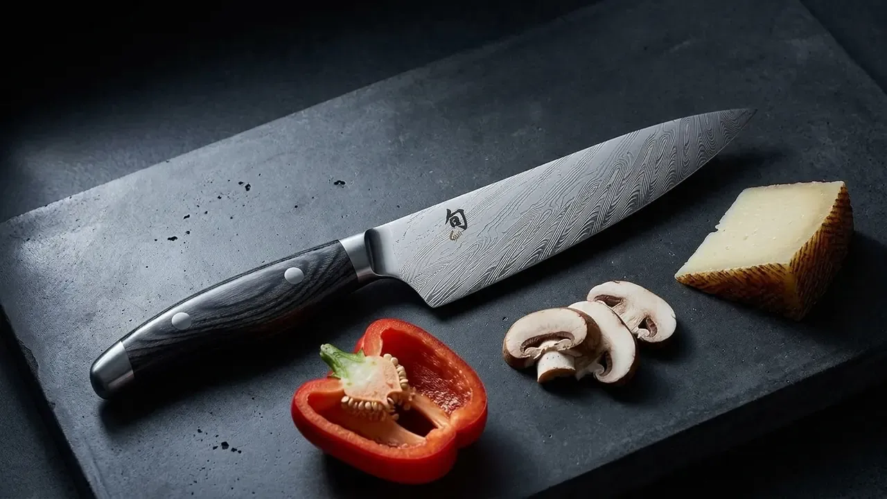 KAI Shun Nagare multiusos de 15 cm con veta de damasco ondulada sobre hormigón oscuro junto a medio pimiento rojo y champiñones laminados
