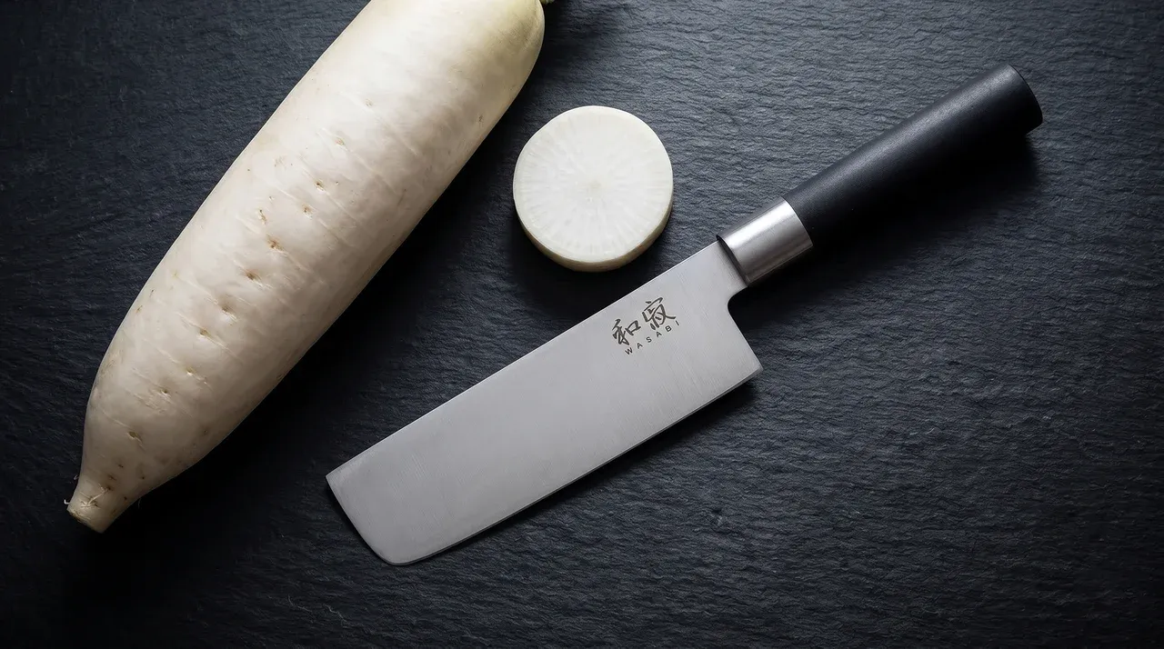 KAI Wasabi Black Nakiri 16,5 cm sobre pizarra oscura con rodajas de daikon