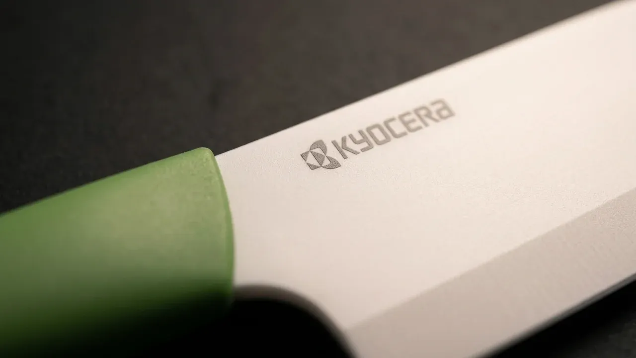 Detalle de la hoja blanca de cerámica de circonio del santoku Kyocera con el logo KYOCERA grabado, junto al mango ergonómico verde