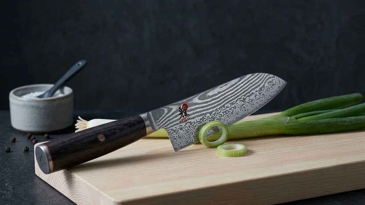 Santoku Miyabi 5000 FCD de hoja damasco y mango de abedul cortando cebolleta sobre una tabla de hinoki, con el logo Miyabi en la hoja