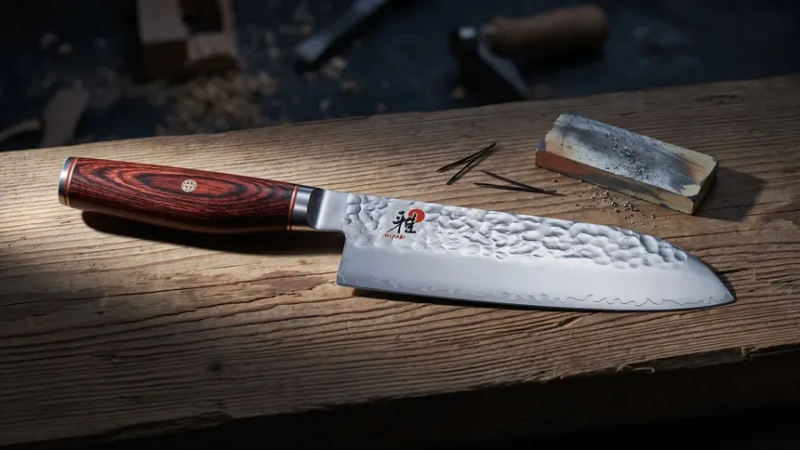 Miyabi 6000MCT Santoku de 18 cm con mango de pakkawood rojizo sobre tablón de hinoki envejecido, junto a una piedra de afilar usada, en un taller de luz fría