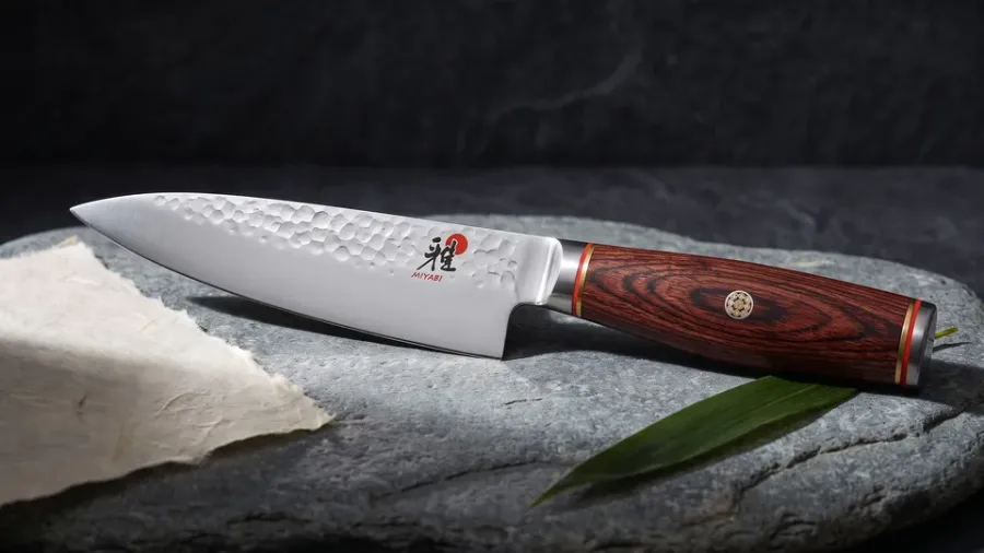 Miyabi 6000MCT Shotoh con mango de pakkawood rojizo sobre losa de piedra de río, junto a un fragmento de washi y una hoja de bambú