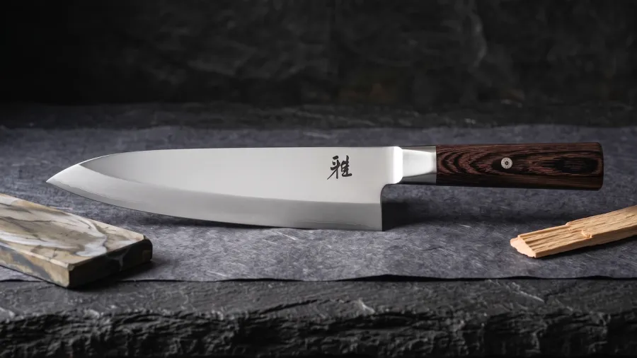 Miyabi KOH 4000FC Gyutoh de 24 cm sobre papel washi oscuro, junto a un fragmento de piedra de afilar y una esquirla de madera de hinoki