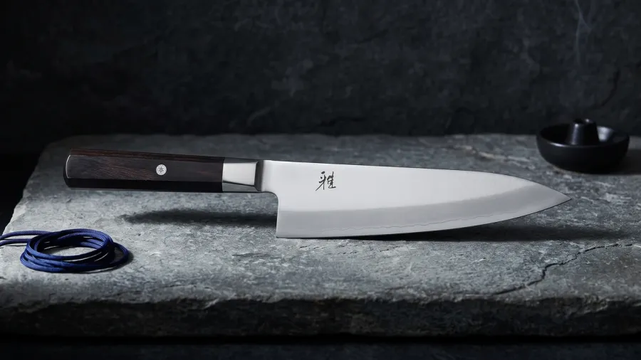 Miyabi KOH Chef de 20 cm sobre losa de piedra de río desgastada, junto a un nudo de cuerda de seda índigo y un pequeño incensario de cerámica negra