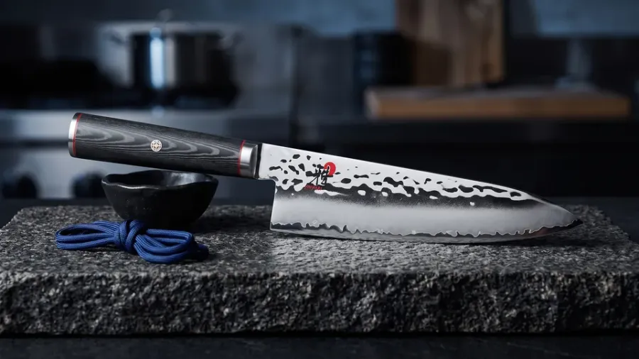 Miyabi Mizu SG2 Chef de 20 cm con mango de micarta negra sobre granito oscuro, junto a un cuenco de cerámica wabi y un cordón de seda índigo