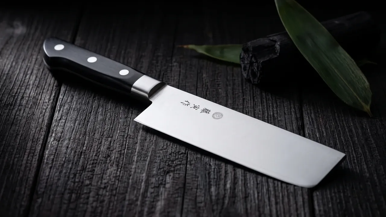 Cuchillo Tojiro Fujitora Usuba de hoja rectangular y filo plano sobre tablón de hinoki claro, junto a un fragmento de carbón y una hoja de bambú, con luz lateral