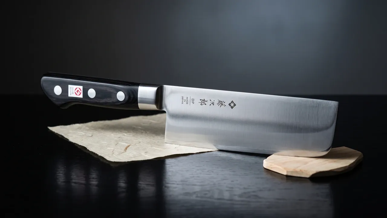 Cuchillo Tojiro 4099 Nakiri de hoja alta y rectangular sobre laca negra, junto a un cuenco wabi y agujas de pino, con luz lateral